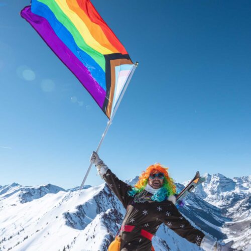 A skier celebrating pride waving a flag and wearing a rainbow wig at Aspen Snowmass in Colorado.