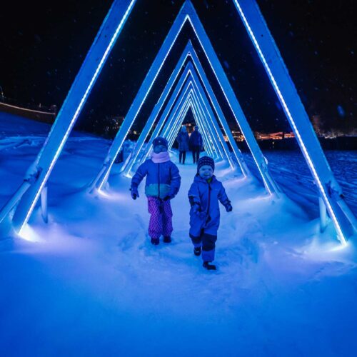 A family walking through the snow under a triangular light tunnel at Aspen Snowmass in Colorado.