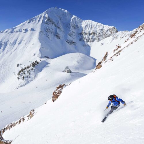 A Mountain Collective skier in a blue jacket skiing a steep slope at Big Sky Resort in Montana.