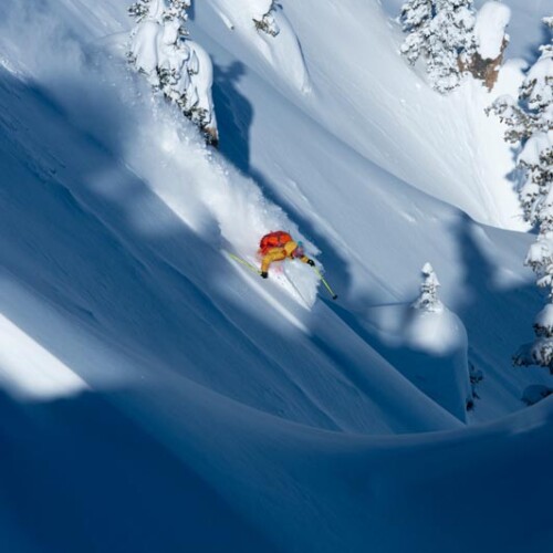 A skier wearing a yellow jacket and red backpack skiing powder at Alta Ski Area with Mountain Collective.