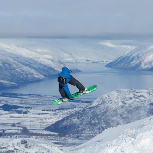 A snowboarder in a blue jacket and green board gets some air at Coronet Peak + The Remarkables.