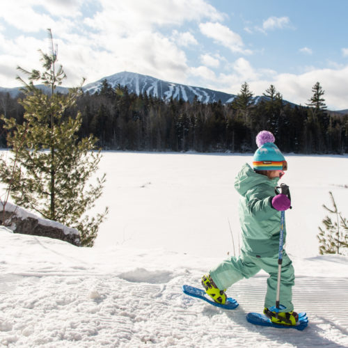 A young child in mint green snow gear snowshoeing at Sugarloaf with the ski mountain in the background.