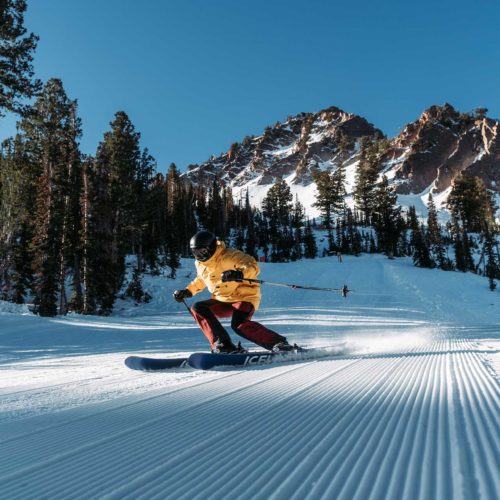 A skier in a yellow jacket carving on corduroy at Snowbasin with Mountain peaks in the background.