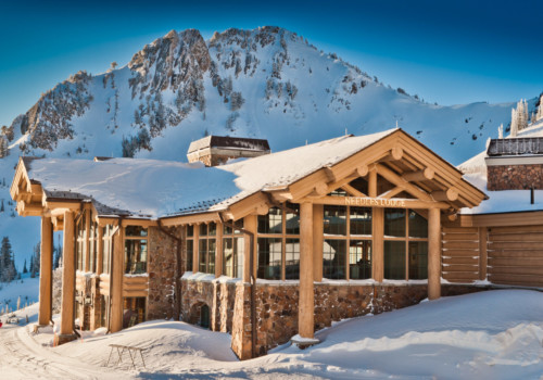 Scenic view of Needles Lodge at Snowbasin Resort, surrounded by snow-covered mountains. The ski lodge features rustic architecture and large windows, offering a cozy retreat in a winter wonderland.