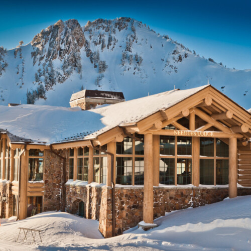 Scenic view of Needles Lodge at Snowbasin Resort, surrounded by snow-covered mountains. The ski lodge features rustic architecture and large windows, offering a cozy retreat in a winter wonderland.