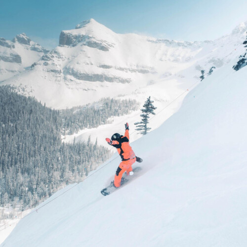 A snowboarder in an orange snowsuit with their arms up going down a mountain