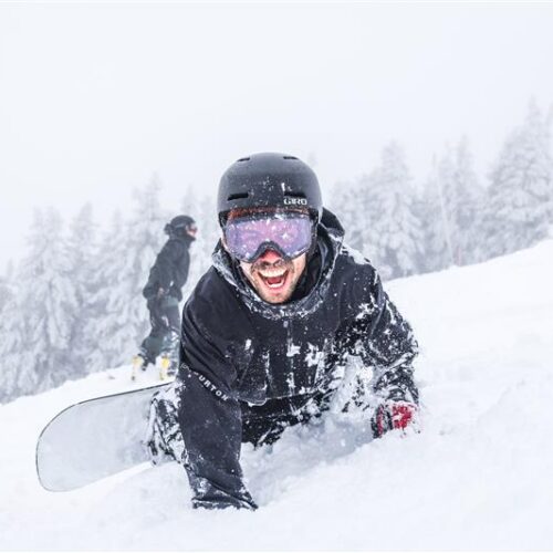 A person wearing a helmet and goggles is excitedly lying on a snowy slope while holding a snowboard. Another person is seen snowboarding in the background, surrounded by snow-covered trees.