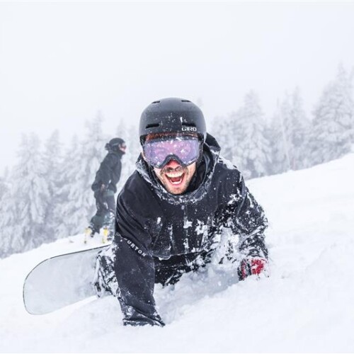 Une personne portant un casque et des lunettes de protection est allongée avec excitation sur une pente enneigée tout en tenant un snowboard. Une autre personne est vue snowboarden arrière-plan, entouré d'arbres enneigés.