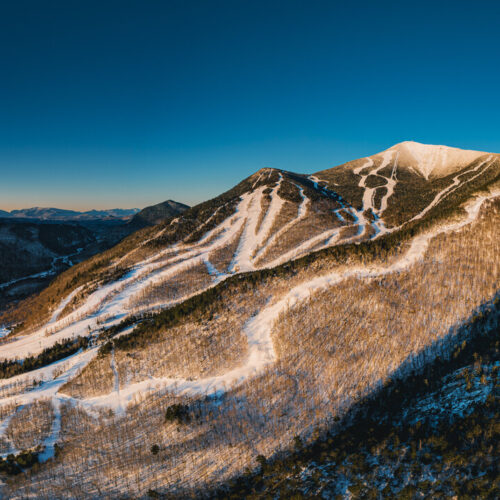 Snow-covered mountain with ski trails illuminated by sunset light, nestled in a forested landscape. The sky is clear and blue, with distant mountain ranges on the horizon.