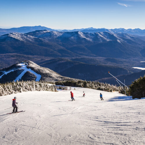 Skiers glide down a snowy slope at Whiteface surrounded by a mountainous landscape. The sky is clear and blue, and tall, forested mountains stretch into the distance under the bright sunlight.