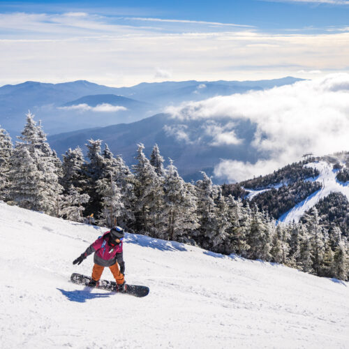 A snowboarder in orange pants and a pink jacket descends a snowy mountain slope, surrounded by snow-covered trees and distant mountainous landscape under a partly cloudy sky.