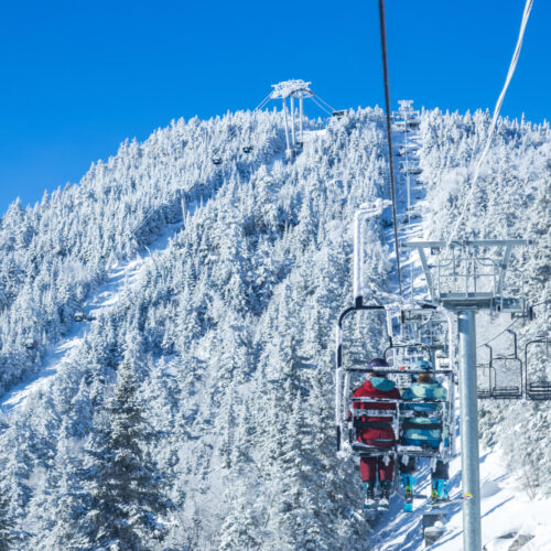 Skiers sit on a chairlift ascending a snowy mountain, surrounded by frost-covered trees under a clear blue sky.