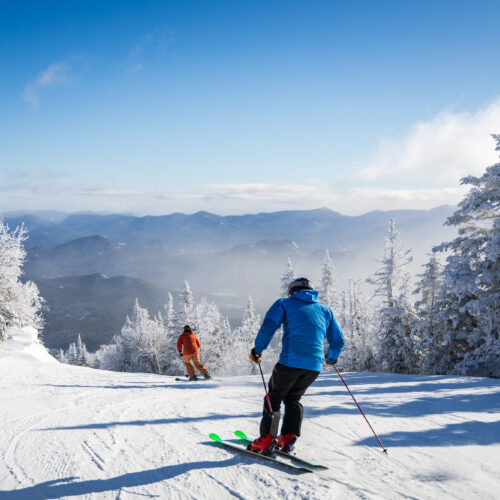 Two people are skiing downhill on a snow-covered mountain slope at Whiteface surrounded by frosted trees, with a clear blue sky and distant mountain range in the background.